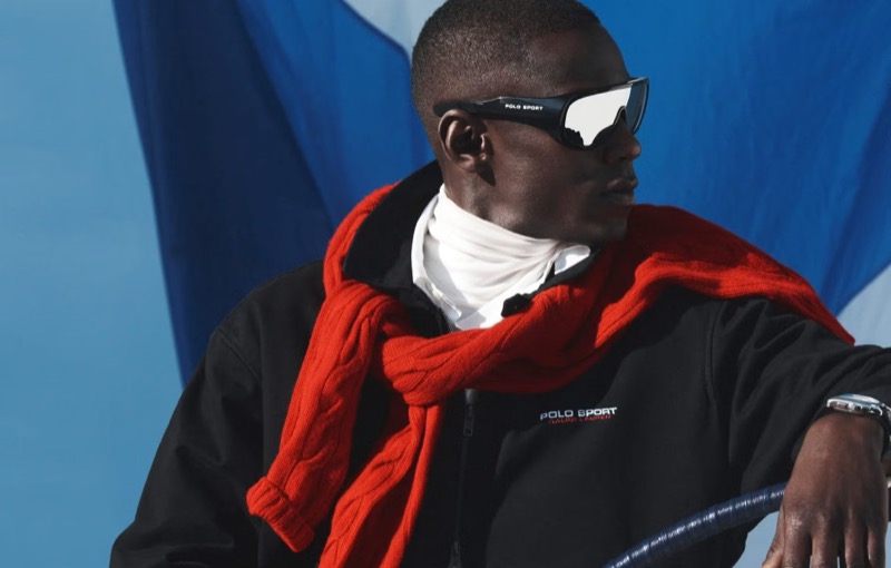 Cheikh wears Polo Sport visor sunglasses, a black Polo Sport zip jacket, a white turtleneck, and a red cable-knit sweater draped across his shoulders, a blue and white Polo Sport flag rising behind him