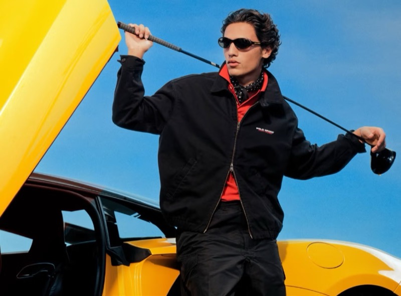 Morgan stands beside a yellow sports car in dark sunglasses, a black Polo Sport zip jacket layered over a red zip polo and black bandana, black trousers, and a golf club lifted across his shoulders