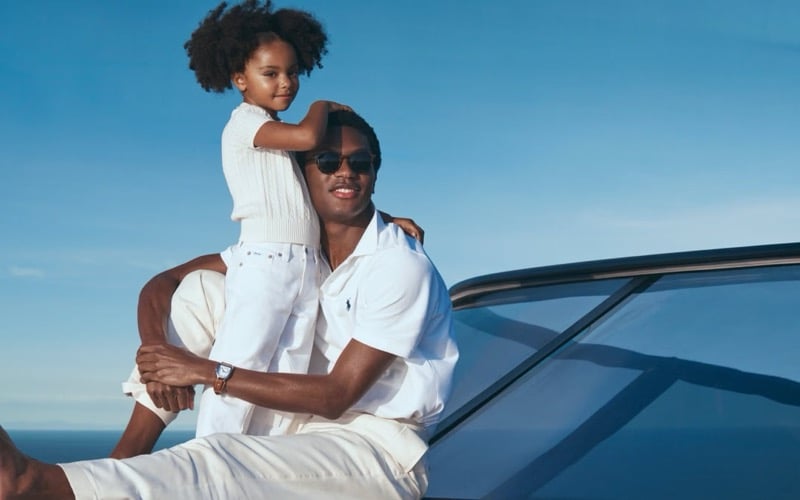 Hamid sits against the coast in sunglasses, a white Polo Ralph Lauren cotton shirt, and white trousers, a small girl in a white cable-knit polo perched on his shoulder with the ocean horizon behind them