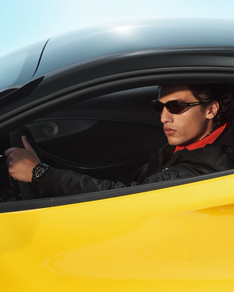 Morgan sits behind the wheel of a yellow sports car in Polo Sport visor sunglasses, a black Polo Sport jacket, and a red zip polo, hand on the steering wheel, the door open to a blue sky