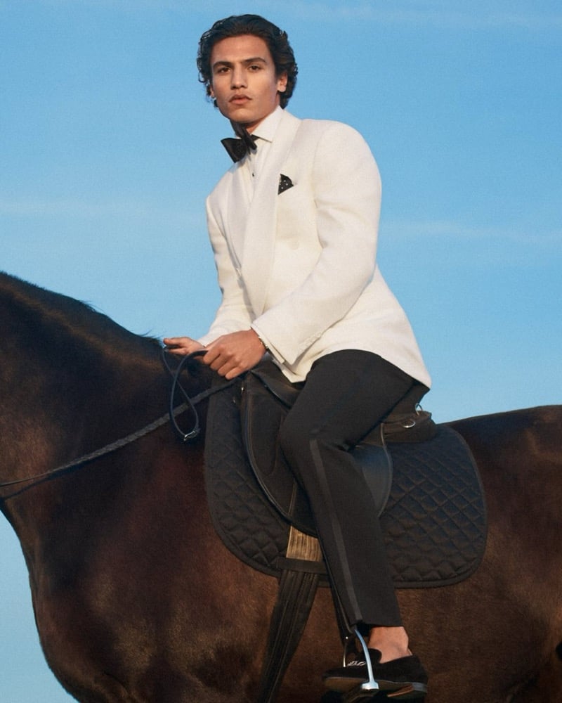 Morgan sits upright on a dark horse against a blue sky in a Purple Label white dinner jacket, black bow tie, white shirt with a black pocket square, black tuxedo trousers with side stripe, and black velvet loafers