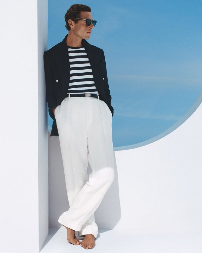Jon Paul leans against a whitewashed wall in sunglasses, a navy double-breasted blazer worn open over a navy-and-white horizontal striped knit, white pleated wide-leg linen trousers, and bare feet, a blue sky behind him