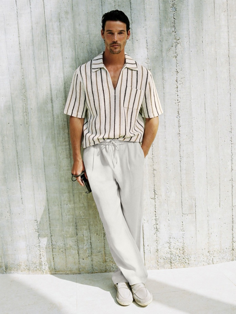 Jorge stands against a concrete wall in the cream Reiss Chatham crochet stripe overhead shirt with brown and tan stripes, white Barges relaxed-fit linen drawstring trousers, and suede penny loafers, holding sunglasses