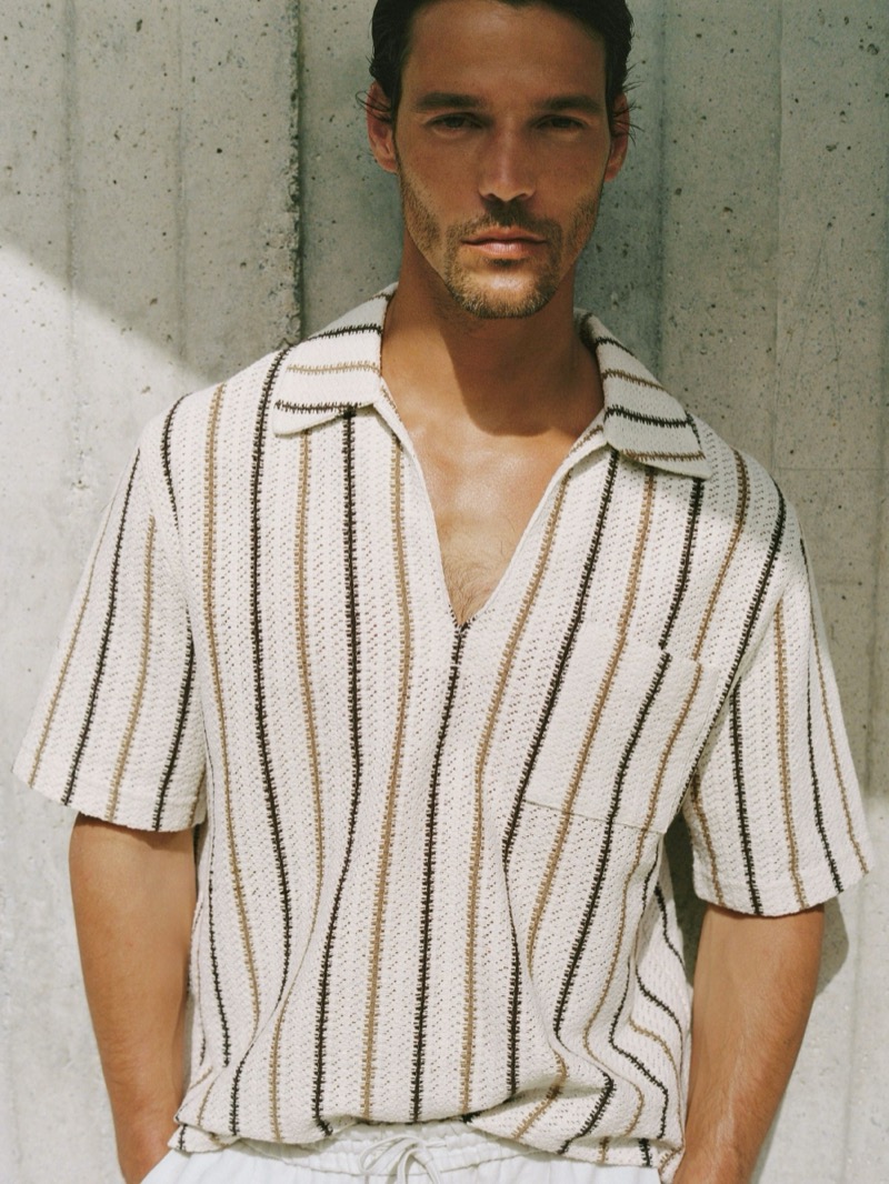 Close-up of Jorge in a cream Reiss Chatham crochet-knit overhead shirt with vertical brown and tan stripes, open at the neck against a concrete backdrop