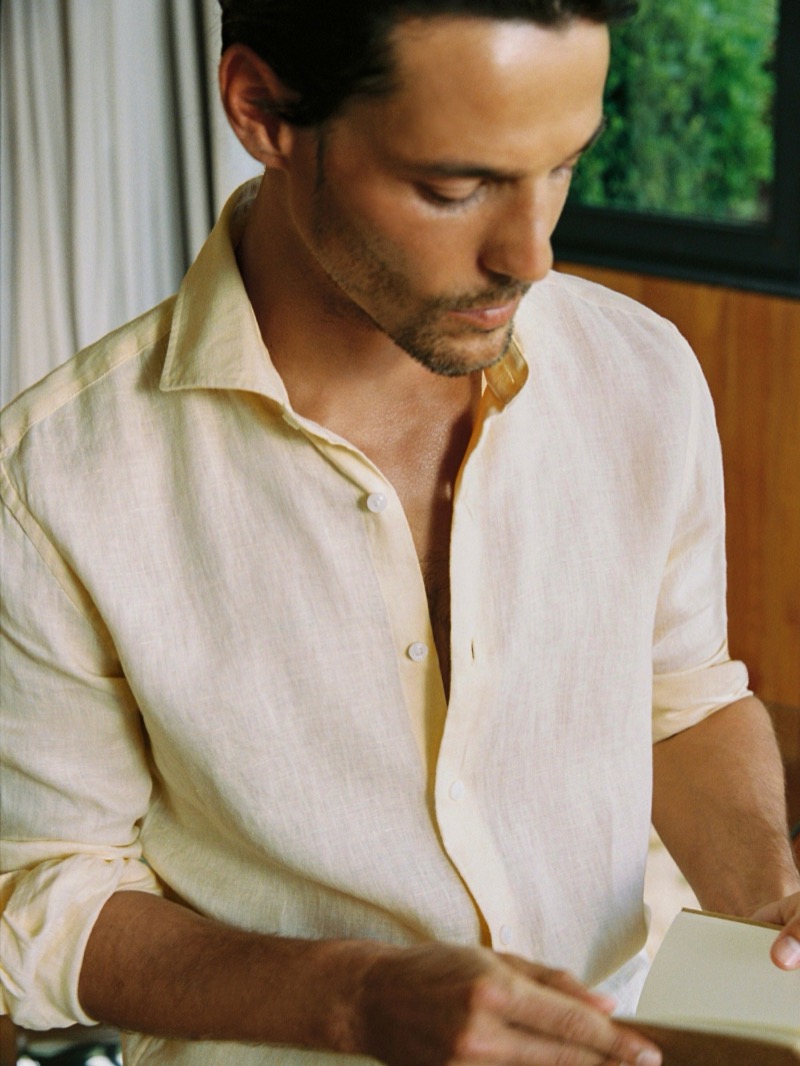 Jorge holds an open book wearing a buttermilk yellow Reiss Ruban linen cutaway-collar shirt with sleeves rolled and open at the neck against bare chest