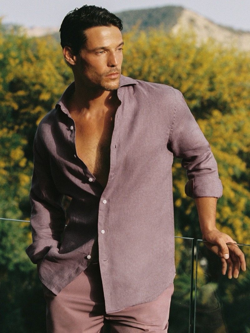 Jorge leans against a glass railing in front of yellow flowering bushes in a mauve pink Reiss Ruban linen shirt with sleeves rolled and worn open at the neck, paired with dusty pink trousers