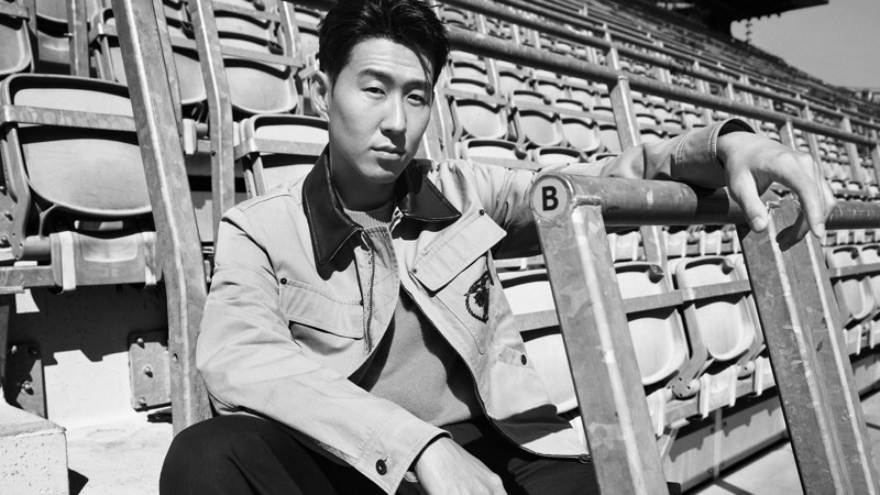 Black and white image of Son crouched at the front of an empty stand of stadium seats, leaning one arm on a railing marked with the letter B, wearing a light tan canvas zip jacket with an Equestrian Knight emblem patched at the chest over a knit crewneck
