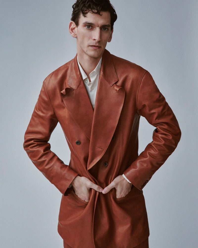 Thibaud Charon in close portrait wearing a Soshiotsuki rust-colored double-breasted leather jacket left open over a white spread-collar shirt, hands tucked into the front pockets, dark curly hair against a neutral gray studio backdrop