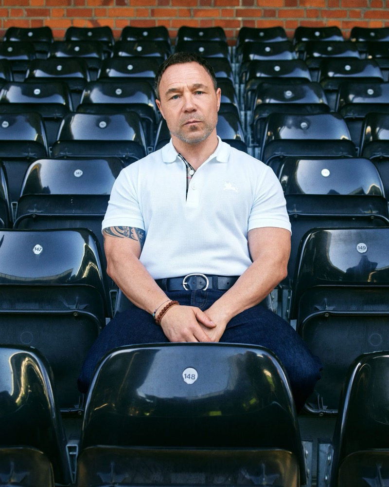 Stephen Graham sits at the front of an empty section of black stadium seats against a red brick wall, wearing a white Burberry pique polo with the Equestrian Knight logo and a Burberry check inner placket, a navy belt, and dark indigo jeans, a forearm tattoo visible