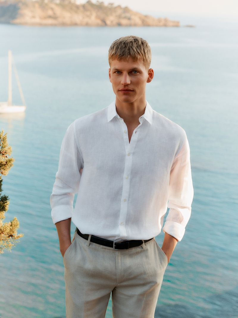 Timo stands by the sea with a sailboat in the distance, wearing a white linen button-down shirt with sleeves rolled and oat linen trousers belted in black