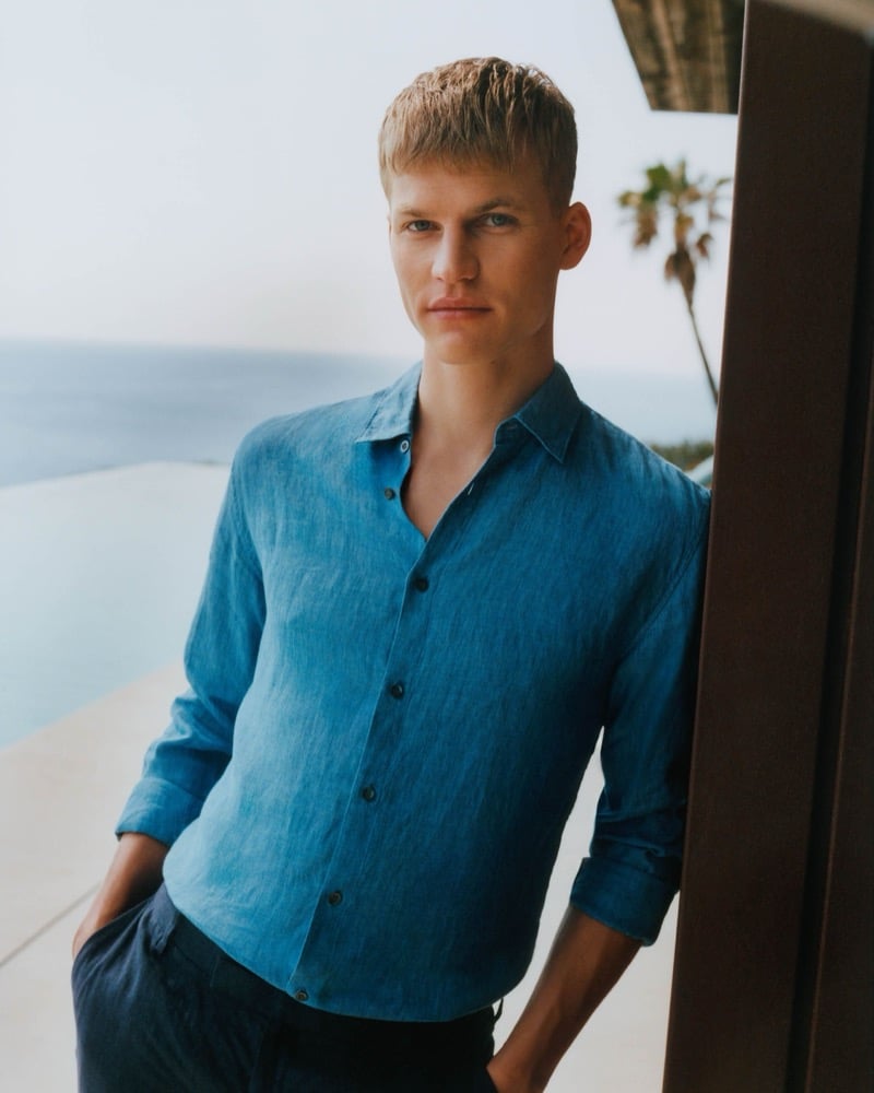 Timo leans against a wood post on a seaside terrace wearing an unbuttoned cool blue linen shirt with navy linen trousers