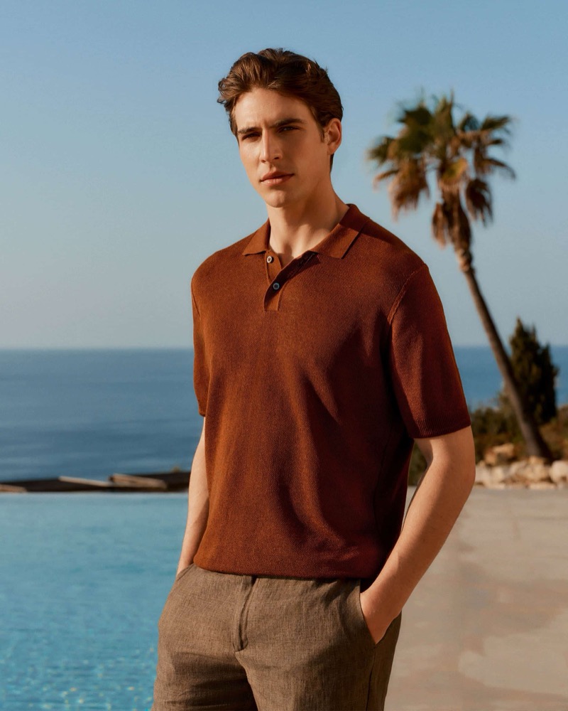Amerigo stands poolside beneath a palm tree in a garnet red short-sleeve knit polo shirt and brown linen trousers