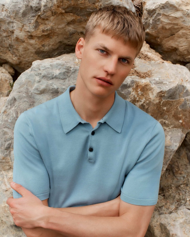 Timo crosses his arms against a pale rock backdrop in a soft denim blue short-sleeve knit polo shirt