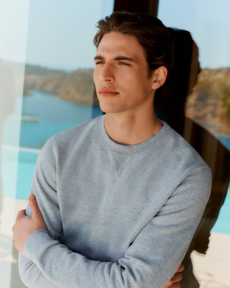 Amerigo stands behind a pane of glass in a heather gray cotton sweatshirt with a coastal cove view behind him