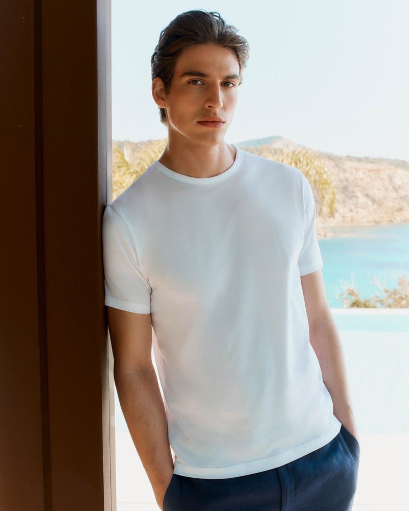 Amerigo leans against a dark wood doorway with a sea view behind him in a white crew-neck cotton T-shirt and navy linen trousers
