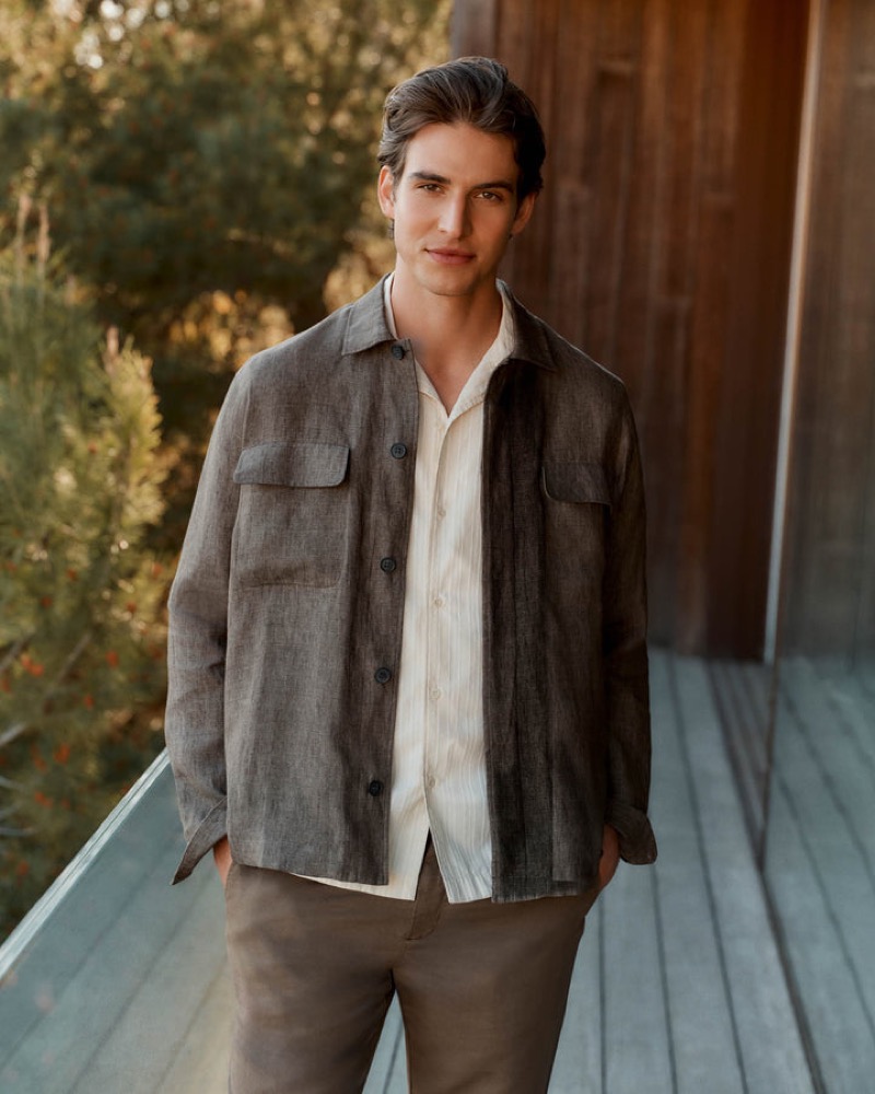 Amerigo stands on a wood deck in a charcoal brown linen overshirt with chest pockets, layered over a cream linen shirt with taupe linen trousers