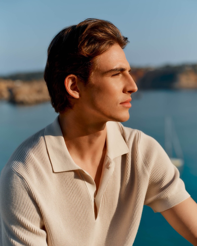 Amerigo looks toward the sea in profile in a cream ribbed knit short-sleeve polo shirt with a coastal cove behind him