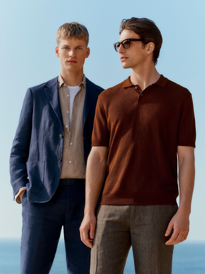 Timo wears a light navy linen blazer over a beige linen shirt and white T-shirt with navy linen trousers beside Amerigo in a garnet red knit polo, brown linen trousers, and tortoise sunglasses