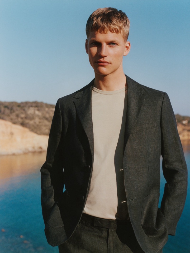 Timo stands by the sea in a charcoal linen blazer and matching linen trousers worn over a cream cotton T-shirt