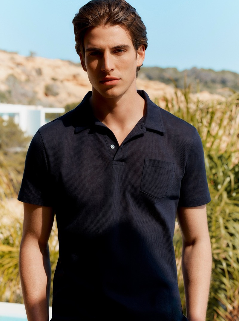 Amerigo stands against coastal grass and rocky terrain in a navy Riviera polo shirt with a chest pocket
