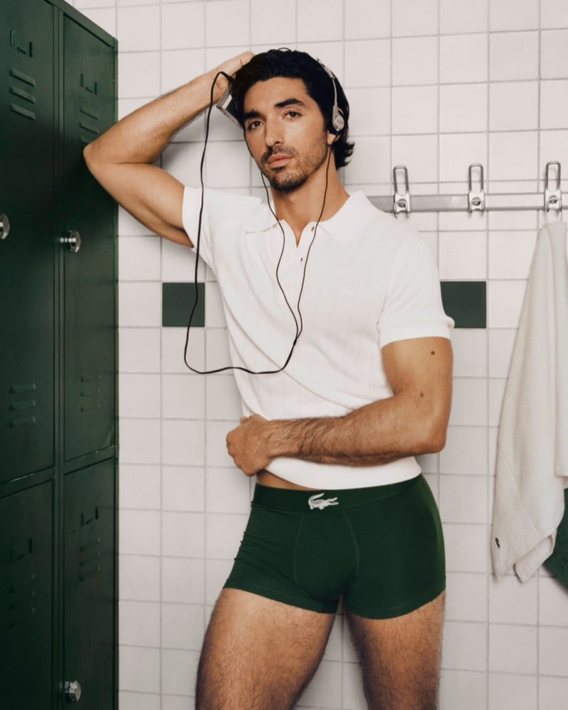 Zakhar Perez stands against a white tile wall beside a green metal locker in a white Lacoste polo and dark green Lacoste boxer trunks with vintage headphones around his neck for the Crocodile Style underwear campaign