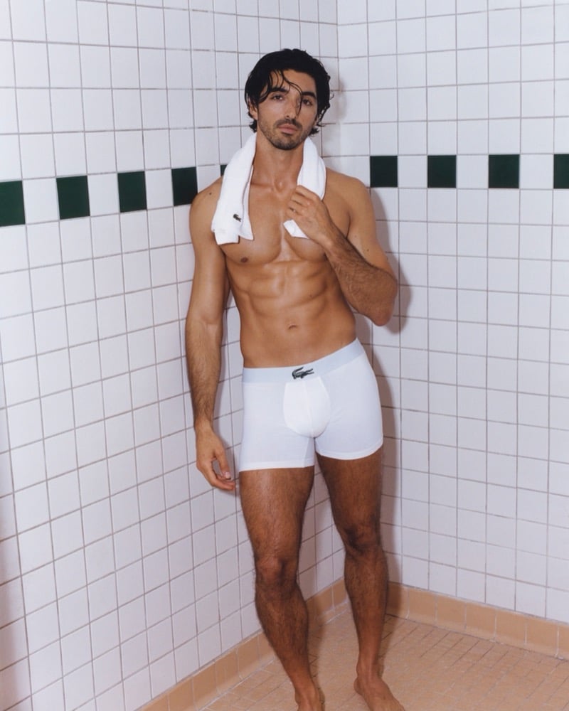 Zakhar Perez stands in a tiled change-room corner with a white Lacoste towel draped around his neck, wearing white Lacoste trunks with the crocodile at the waistband for the Crocodile Style underwear campaign