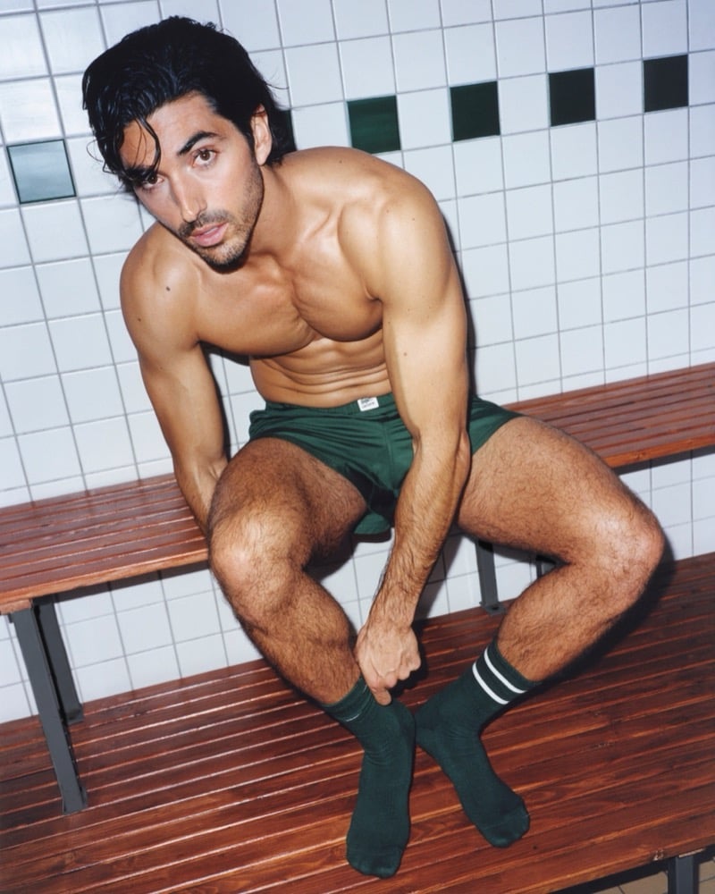 Zakhar Perez sits forward on a wooden bench in dark green Lacoste boxer shorts and dark green athletic socks with white stripes against a tiled change-room wall for the Crocodile Style underwear campaign
