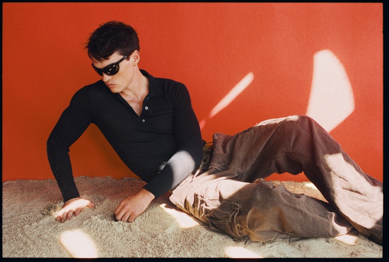 Gabriel Jayne reclines on sandy ground against a deep red wall, wearing black wraparound sunglasses, a long-sleeve black polo, and washed brown cargo pants