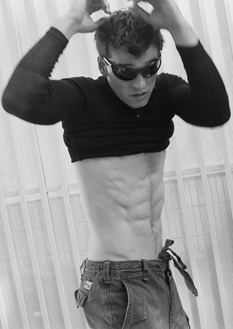 Black and white shot of Gabriel Jayne lifting a black long-sleeve top to reveal his torso, wearing wraparound sunglasses and washed cargo shorts