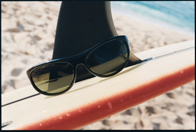 Black Thistles sunglasses with dark lenses rest on the rail of a white and orange surfboard against a sandy beach