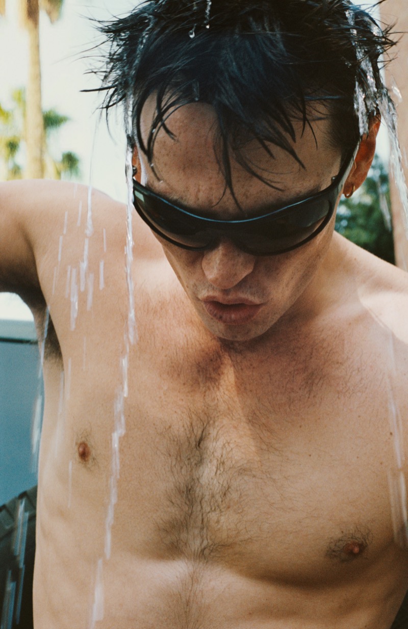 Gabriel Jayne lets water pour over his dark hair while wearing low black wraparound sunglasses, with palm trees in the background