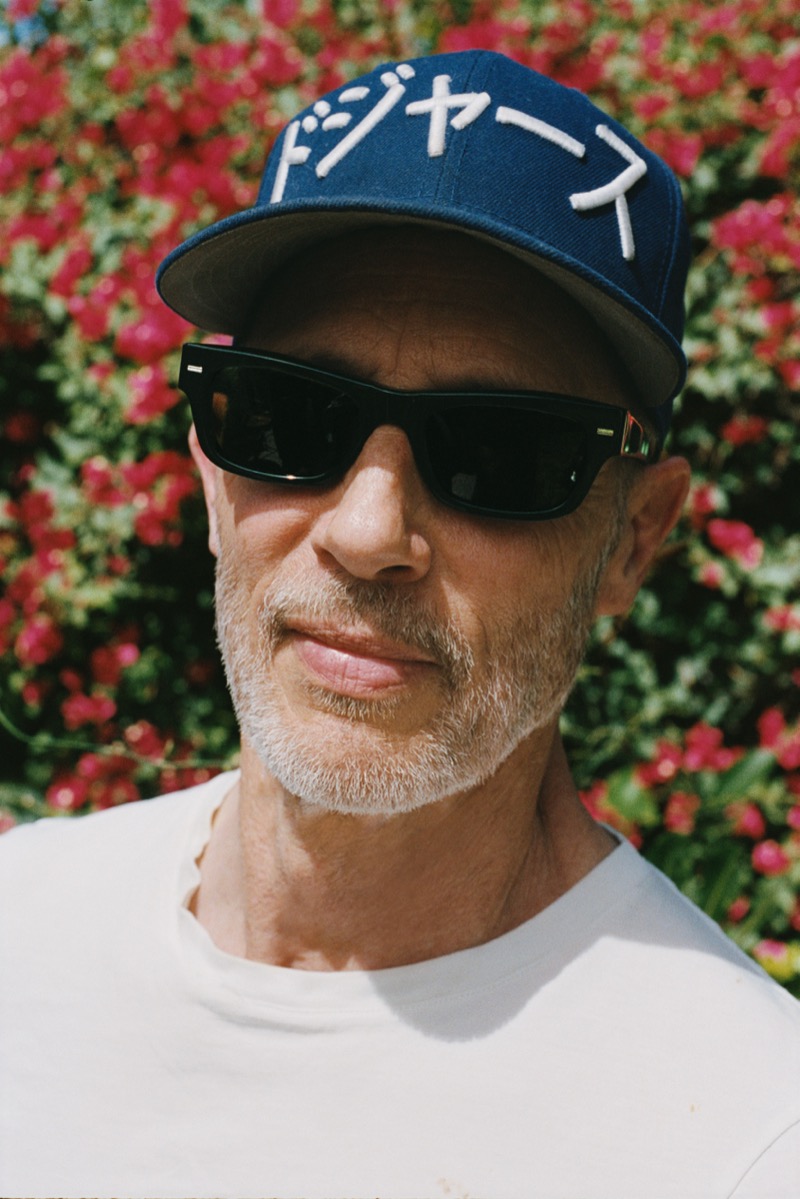 Gries wears a navy Dodgers cap with Japanese lettering, black square sunglasses, and a white T-shirt against a wall of red bougainvillea