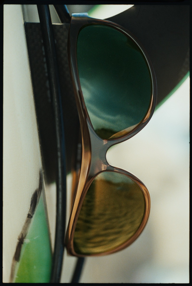 Close-up of a pair of translucent brown Thistles sunglasses with green-tinted lenses, hooked over the edge of a surfboard