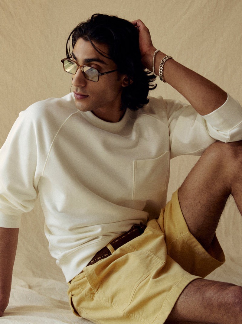 Adarsh sits in a Todd Snyder Foundation pocket sweatshirt in bisque with mustard yellow shorts, a brown leather belt, angular tinted sunglasses, and silver bracelets