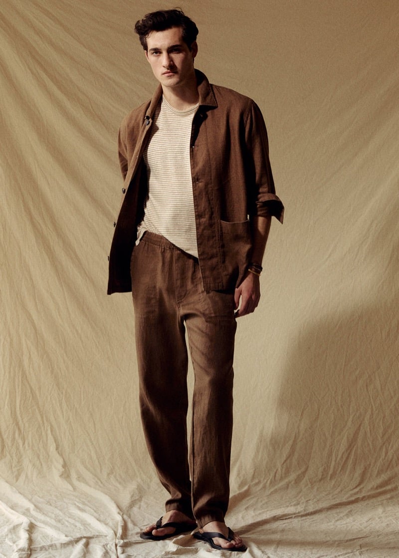 Adam stands in a brown Todd Snyder linen chore coat over a cream and tan striped tee with brown linen beach pants and black thong sandals