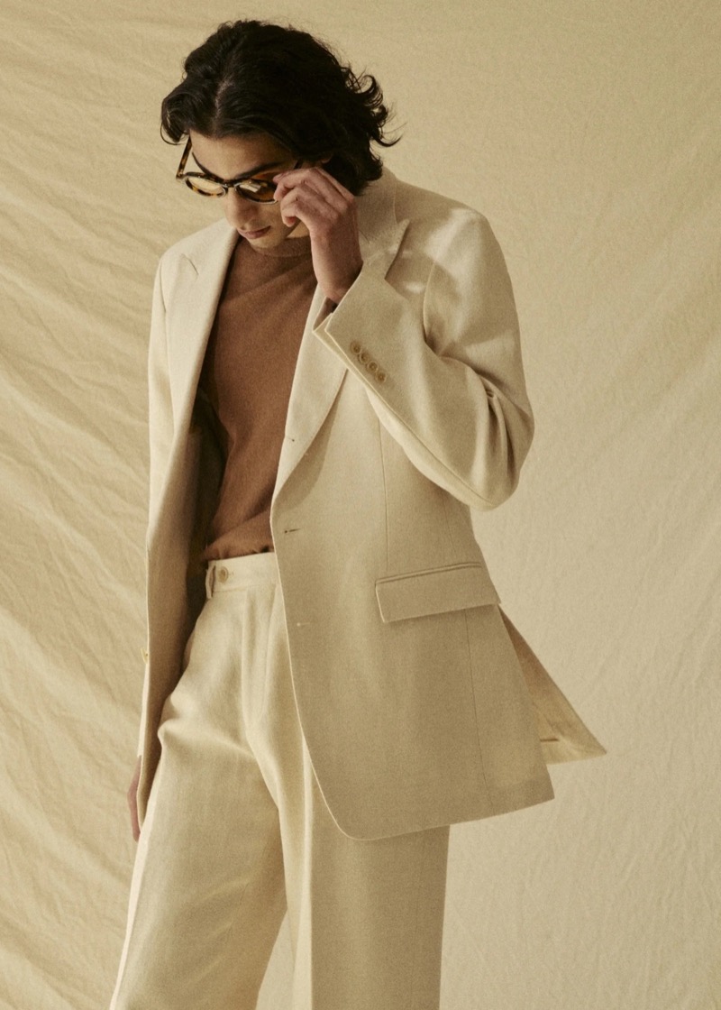 Adarsh wears a Todd Snyder peak-lapel linen Madison jacket in birch over a brown crewneck sweater with matching birch wide-leg trousers and tortoise glasses