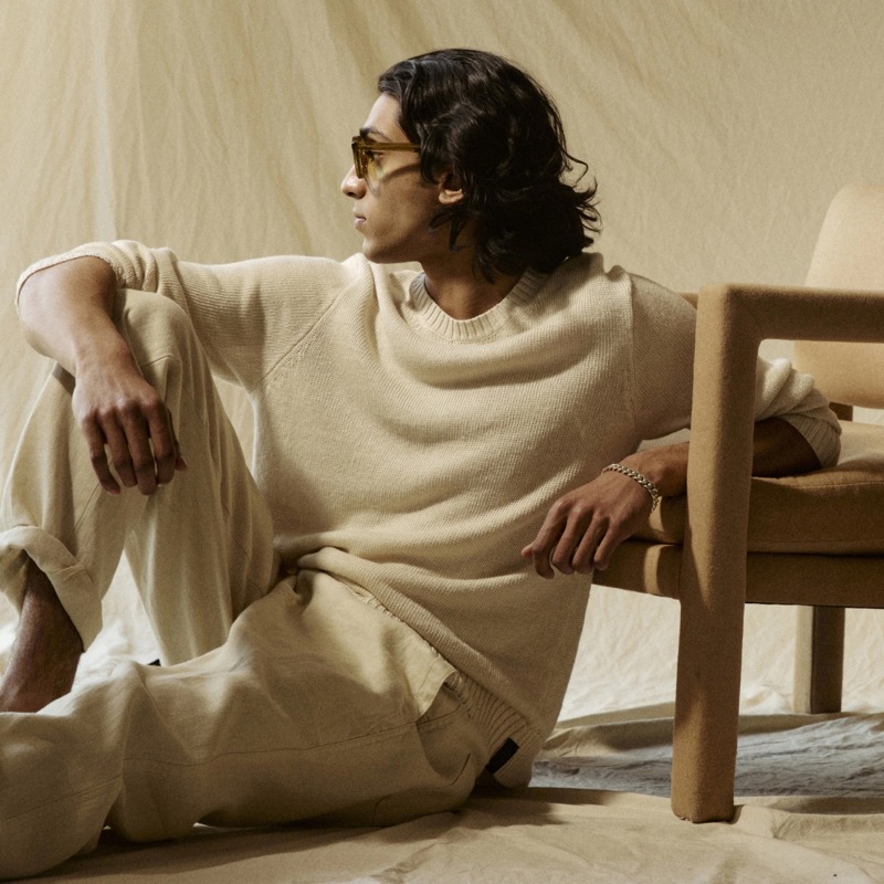 Adarsh sits on the floor in a Todd Snyder bisque linen crewneck sweater with matching bisque trousers, tinted sunglasses, and a silver chain bracelet