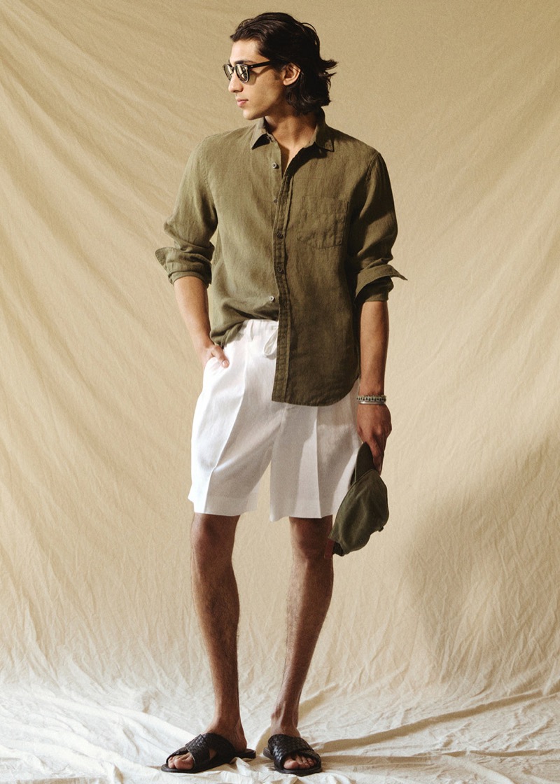Adarsh wears an olive linen button-up shirt with sleeves rolled, white Todd Snyder Mylos pleated linen shorts, black slide sandals, and holds an olive cap