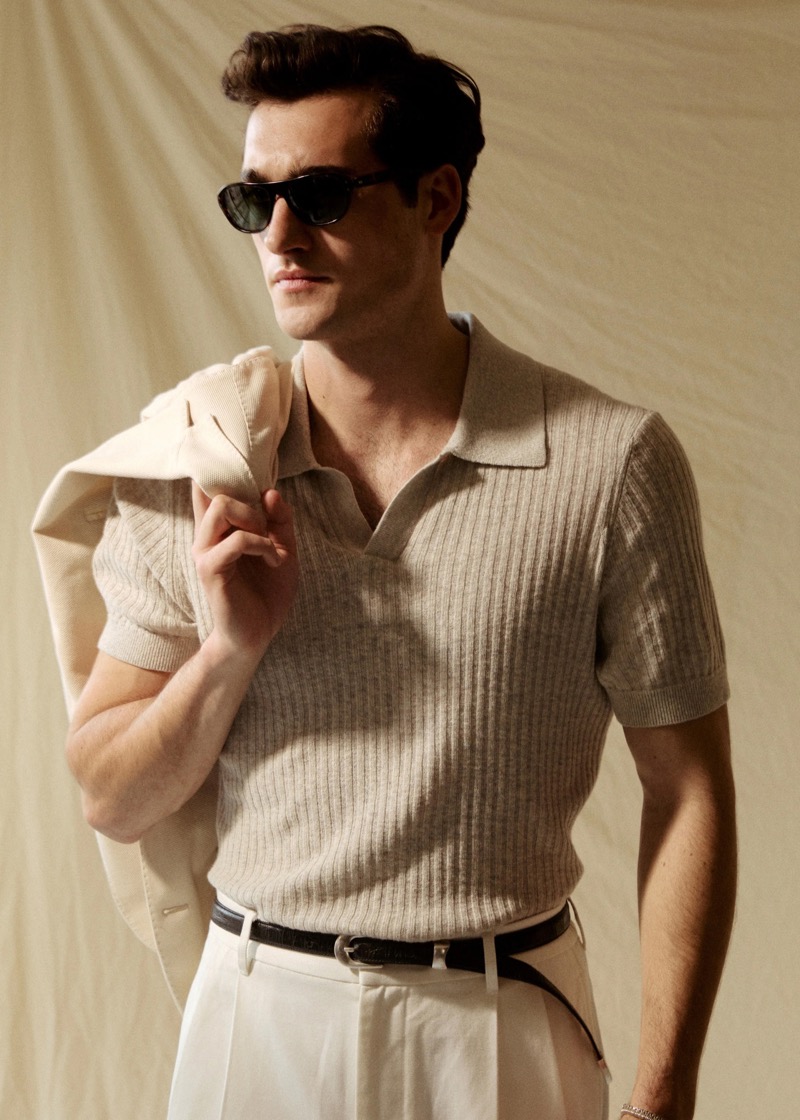 Adam wears a Todd Snyder Tide Montauk ribbed knit polo in dusty steel with a black belt and cream pleated trousers, holding a tan jacket over one shoulder