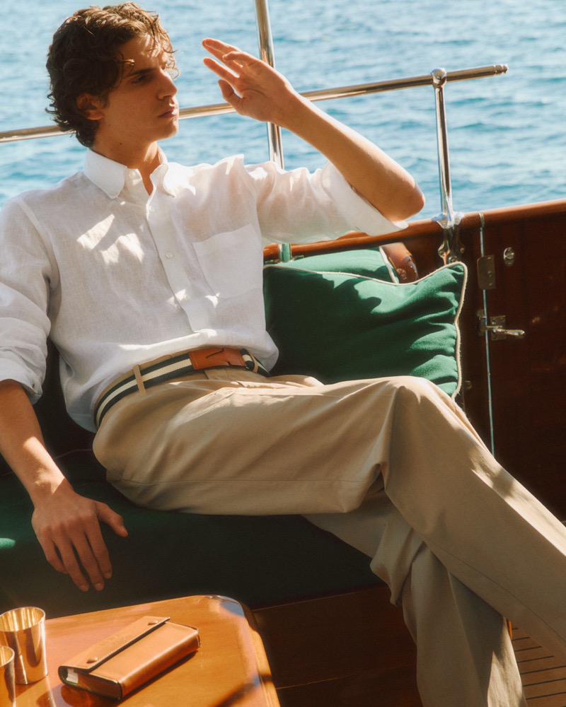 Hedi reclines on a forest green deck cushion shading his eyes, in a white linen button-down shirt and stone chinos belted with a striped green, cream, and black Greca webbing belt