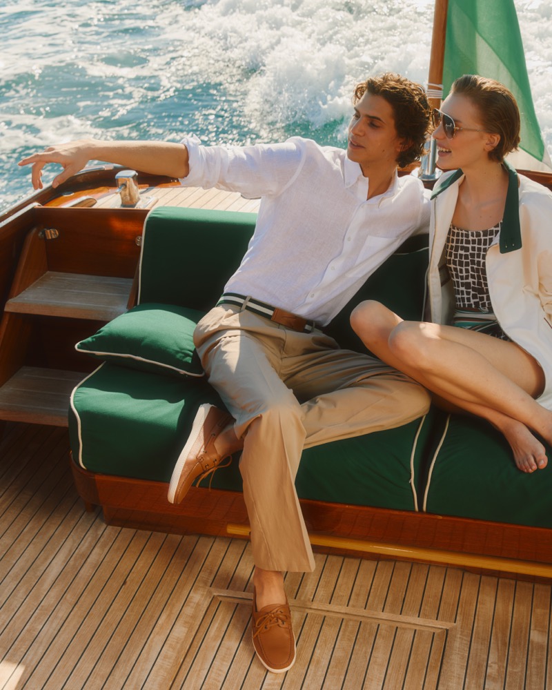 Hedi sits on a forest green deck cushion in a white linen shirt with khaki trousers and tan suede Marlin loafers, beside a female model in a white overshirt and patterned swimsuit