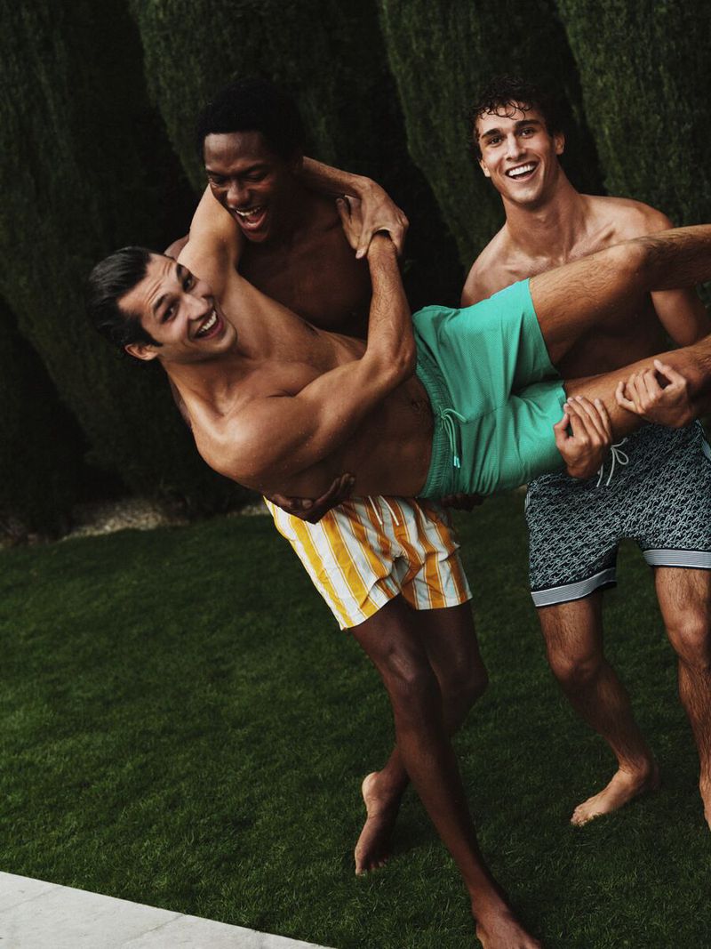 Three friends roughhouse shirtless on a lawn beside the pool, one lifted horizontally in yellow vertical stripe swim shorts, another in mint green mesh trunks, the third in navy diamond-print shorts with a striped hem, tall cypress hedges behind