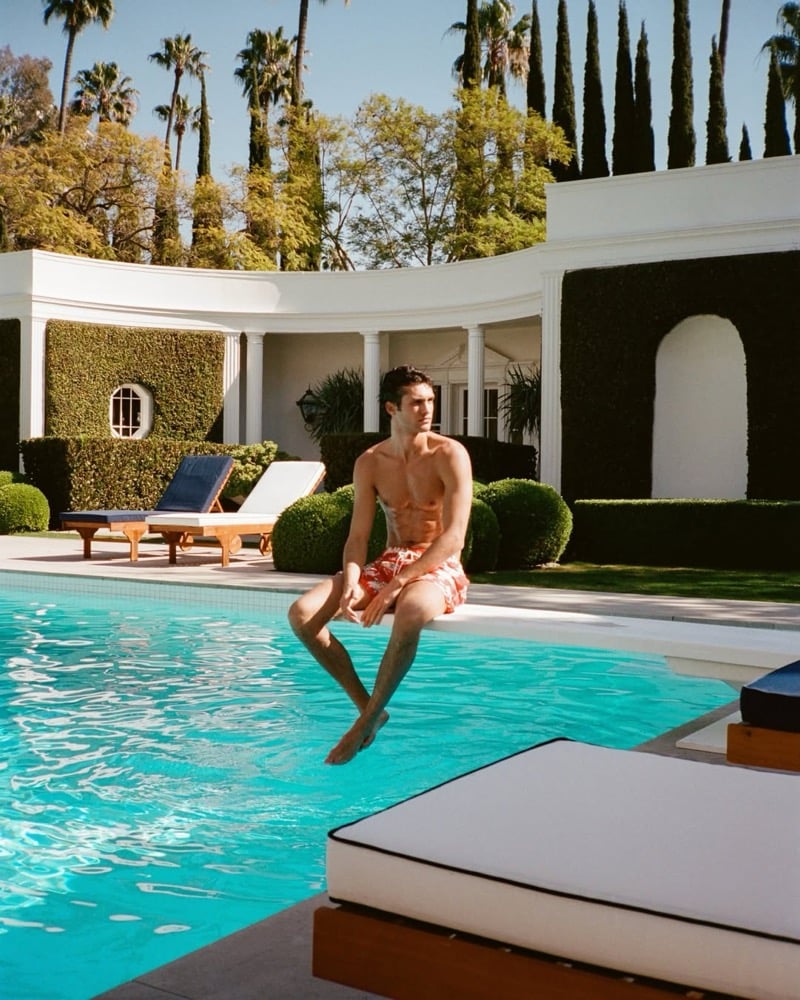Francisco Henriques perched on the pool diving board in coral red printed swim shorts, a vintage Beverly Hills estate of cypress hedges, white columns, and palm trees spread behind him