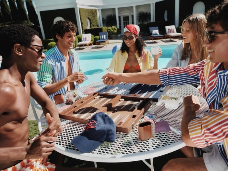 Group of friends gather at an outdoor table beside the pool for a game of backgammon, dressed in red, blue, and yellow vertical stripe resort shirts with a navy Tommy Hilfiger flag cap on the table