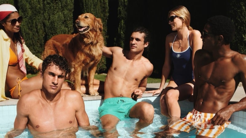 Friends linger at the pool edge with a golden retriever, the men shirtless in mint and yellow stripe swim shorts, the women in a navy one-piece and an orange bikini