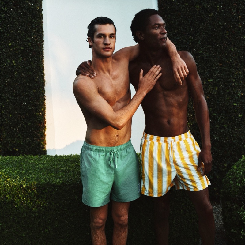 Francisco and Hamid stand arm-in-arm against tall cypress hedges at sunset, one in mint green swim shorts, the other in yellow and white vertical stripe trunks