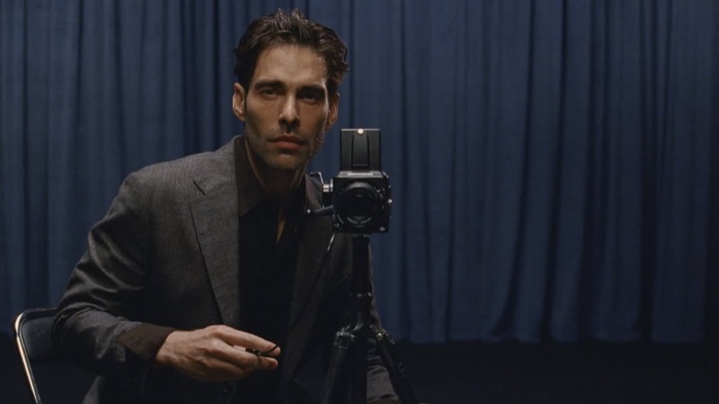 Jon sits behind a medium-format Hasselblad camera on a tripod, wearing a gray textured blazer against a navy curtain