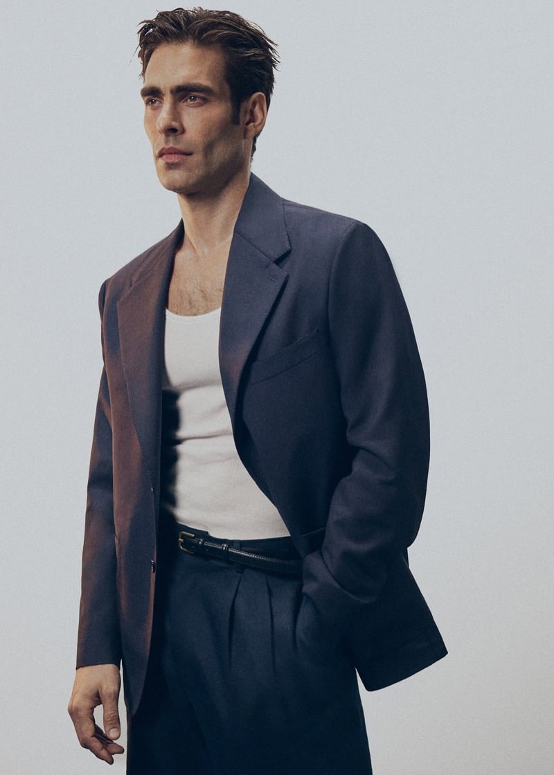 Jon wears a navy unstructured blazer with peak lapels over a white tank top, with navy pleated trousers and a black leather belt