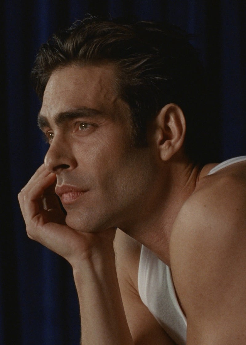 Close-up of Jon in a white cotton tank top against a deep blue curtain, resting his chin on his hand