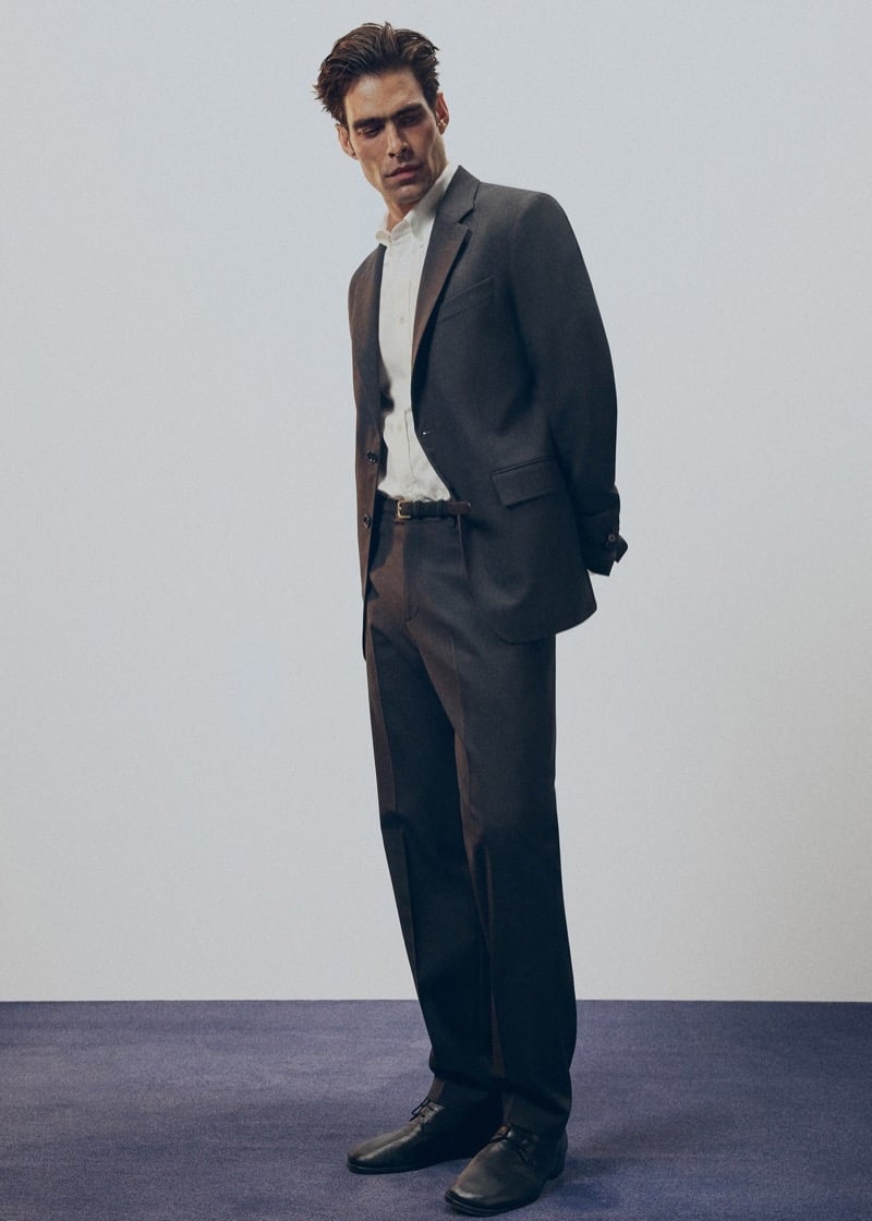 Jon stands in a charcoal gray two-button suit with a white open-collar shirt, brown belt, and dark brown leather shoes